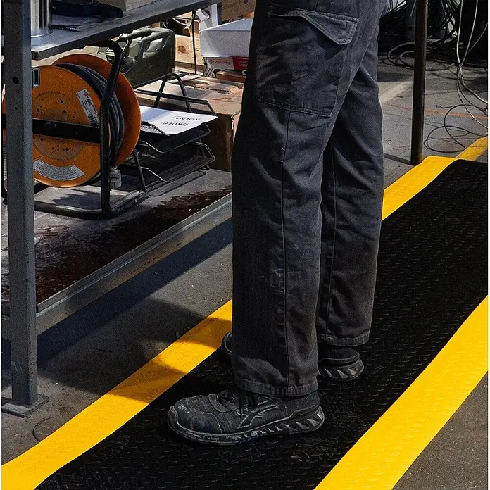 Proregal Gummimatte Anti-Ermüdungsmatte COMFORT PLUS 122x200cm Schwarz/Gelb - 200 x 122 cm, Schwarz/Gelb - Arbeiter steht auf schwarzer Anti-Ermüdungsmatte mit gelben Sicherheitsstreifen im Industriebereich.