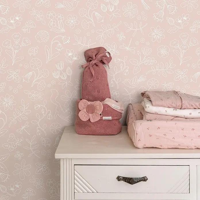 Little Dutch Vliestapete Feentraum - Rosa/Weiß, Floral, 10 x 0,53 m - Rosa gestrickter Babysack mit Blumendetail und gefaltete Textilien auf einer Kommode vor gemusterter Tapete.
