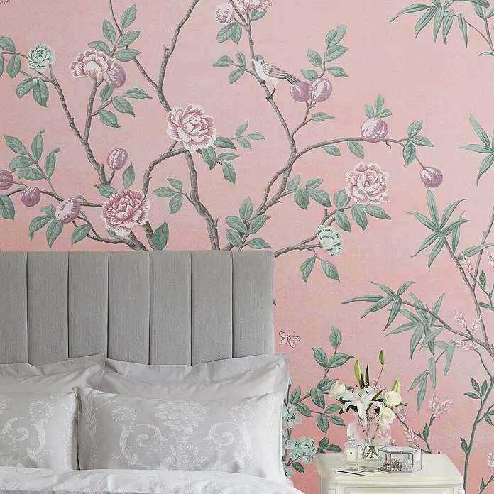 Laura Ashley Fototapete Eglantine - Vlies - Rosafarbene Tapete mit detailliertem Chinoiserie-Muster aus Ästen, Blumen, Früchten und einem Vogel, hinter einem grauen Kopfteil im Schlafzimmer.
