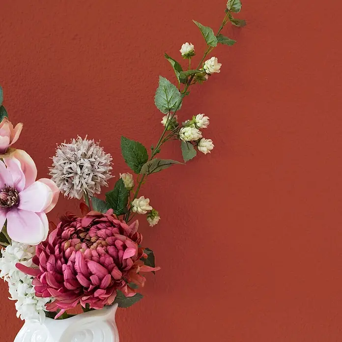 Bricoflor Wandfarbe 2,5 Liter, Merlot - dunkel-rot, Matt, 2,5 l, Wasserverdünnbar - Kunstblumenarrangement mit rosa und weißen Blüten in einer weißen Vase, vor einer strukturierten Terrakottawand.