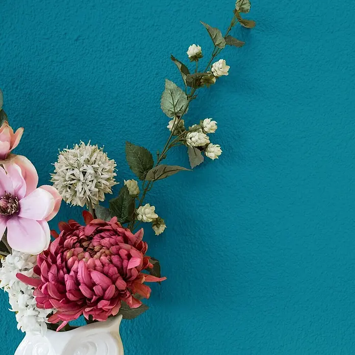 Bricoflor Wandfarbe 2,5 Liter, Petrol - blau grün, Matt, 2,5 l, Wasserverdünnbar - Eine Kunstblumenarrangement mit rosa und weißen Blüten und grünen Blättern in einer weißen Keramikvase, vor einer strukturierten tealfarbenen Wand.