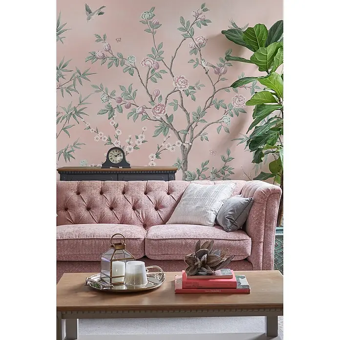 Laura Ashley Fototapete Eglantine - Vlies - Ein rosafarbenes getuftetes Sofa mit Kissen in einem gemütlichen Wohnzimmer vor einer geblümten Tapete.