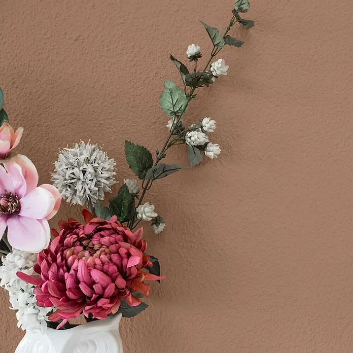 Bricoflor Wandfarbe 2,5 Liter, Lehm - hell-braun, Matt, 2,5 l, Wasserverdünnbar - Ein lebhaftes Bouquet aus rosa, bordeauxroten und weißen Kunstblumen mit grünen Blättern in einer weißen Vase vor einer strukturierten braunen Wand.