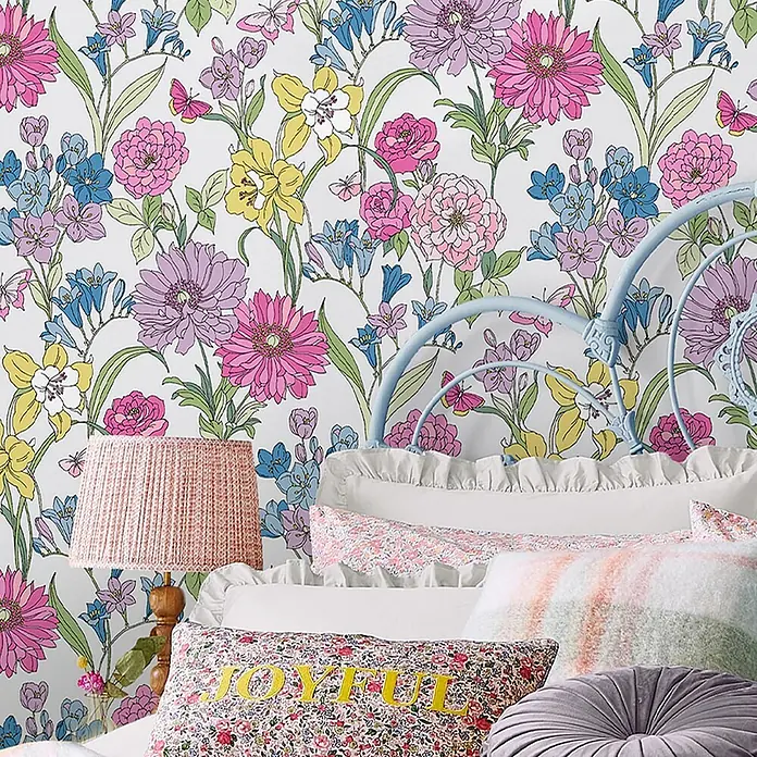 Vliestapete Gilly 1000x52cm Laura Ashley -  - Bunte florale Tapete mit rosa, gelben, blauen und lila Blumen sowie Schmetterlingen schmückt ein Schlafzimmer hinter einem hellblauen Bett und Kissen.
