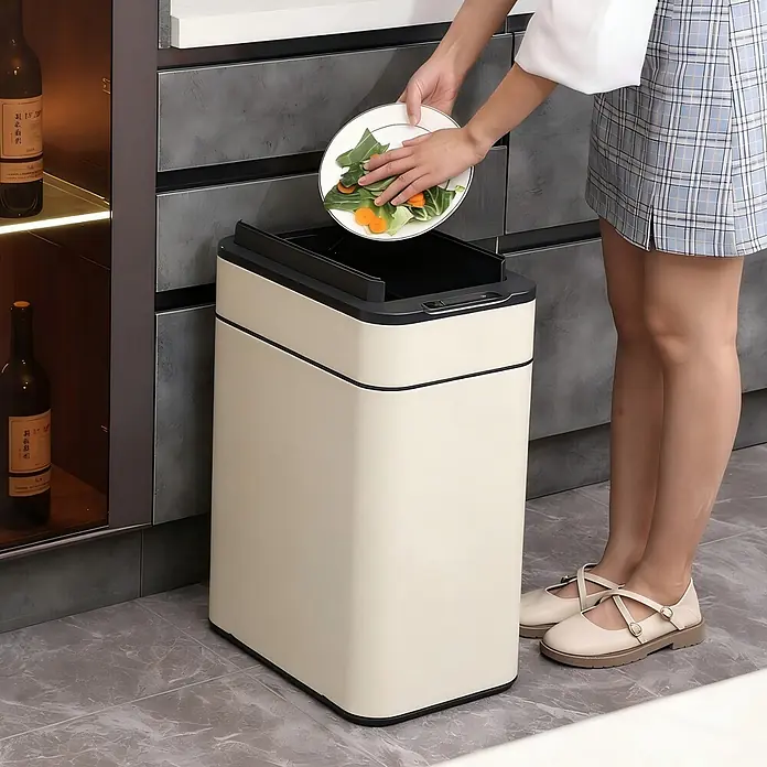 SVITA Sensormülleimer SE50L - 50 l, Stahl, Taupe - Cremefarbener rechteckiger smarter Mülleimer mit schwarzem Deckel, in einer Küche, während eine Person Lebensmittelabfälle hineinwirft.