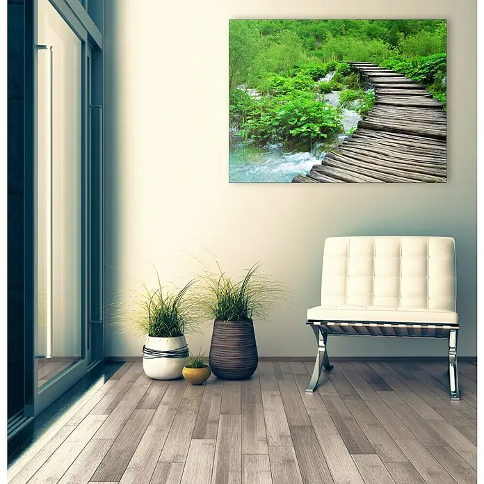 Leinwandbild Young Living - Sparkling Nature, B x H: 120 x 90 cm - Ein modernes Wohnzimmer mit einem stilvollen Barcelona-Stuhl, Pflanzen und einem Wandbild einer Holzbrücke über einen Bach. Helle und natürliche Atmosphäre.