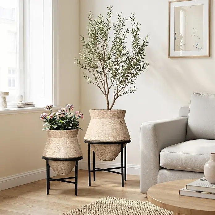 Hometopia Pflanztopf HTPL0359-BR - 2 Stk., Außenmaß (Ø x H): 62,5 x 62,5 cm, Metall, braun - Zwei strukturierte beigefarbene Pflanzgefäße auf schwarzen Metallständern, eines mit rosa Blumen, das andere mit einem Olivenbaum, in einem hellen Wohnzimmer.