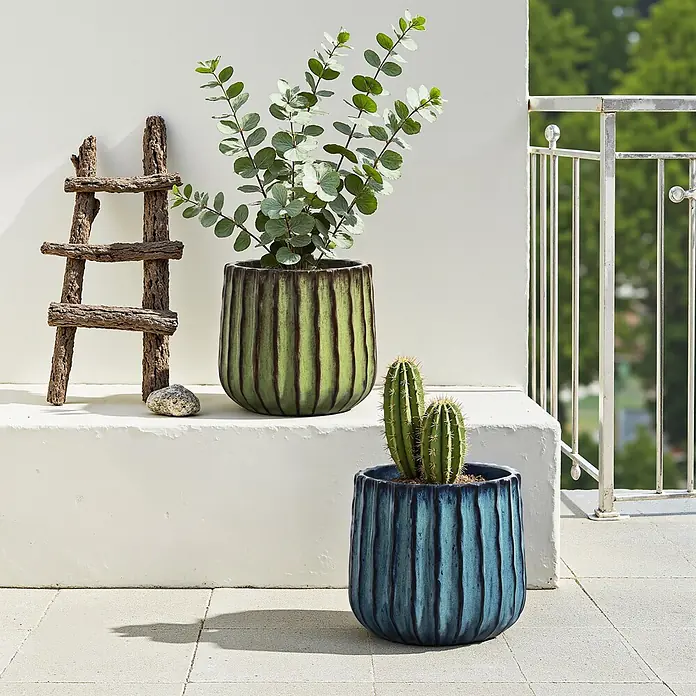 Keramik Blumentopf -  - Zwei gerippte Pflanzgefäße auf einer Terrasse: ein großes grünes mit einer Blattpflanze und ein kleineres blaues mit einem Kaktus.