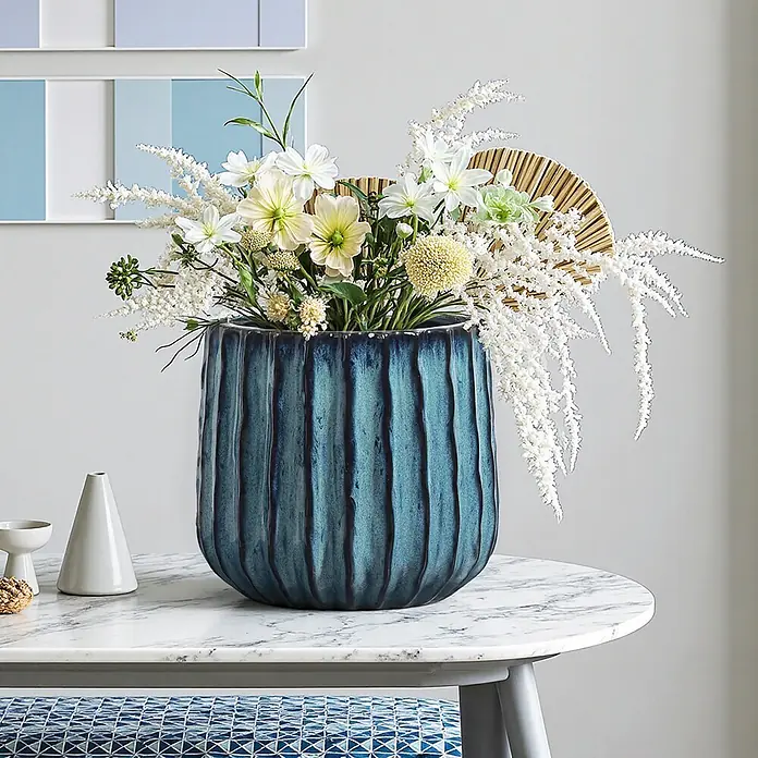 Keramik Blumentopf -  - Eine strukturierte blaue Keramikvase enthält ein lebendiges Arrangement aus weißen und hellgelben Blumen auf einem Marmortisch.
