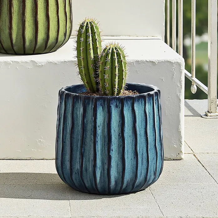 Keramik Blumentopf -  - Ein gerippter, glasierter blauer Pflanztopf mit einem Kaktus auf einer Außenterrasse. Ein teilweise sichtbarer grüner, gerippter Pflanztopf ist im Hintergrund.