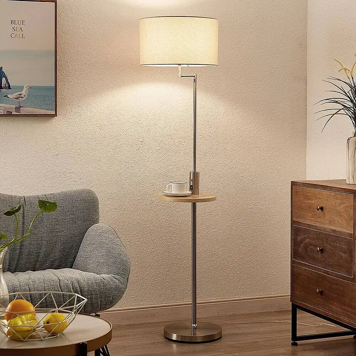 Lindby Stehlampe Wohnzimmer in nickel satiniert, beige, Stehleuchte -  - Stehleuchte mit beigem Stoffschirm, Metallgestell und integriertem Holzregal, das eine Kaffeetasse hält, in einem Wohnzimmer.