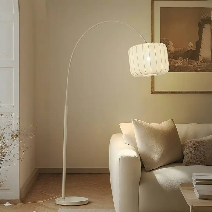 Lindby Stehleuchte Helin - creme / amber, creme / amber, E27 - Große, gebogene Stehlampe in Cremefarbe mit strukturiertem Stoffschirm, die ein warmes Licht auf ein cremefarbenes Sofa in einem Wohnzimmer wirft.