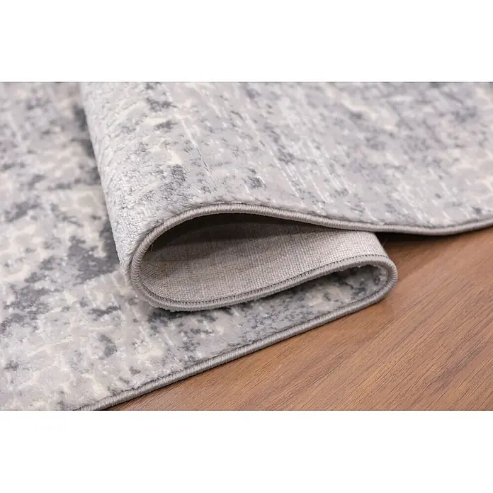 Alfombra de pelo corto Piel - Gris, 230 x 160 cm, 100% poliéster - Alfombra gris claro con patrón abstracto, parcialmente enrollada, sobre un suelo de madera.