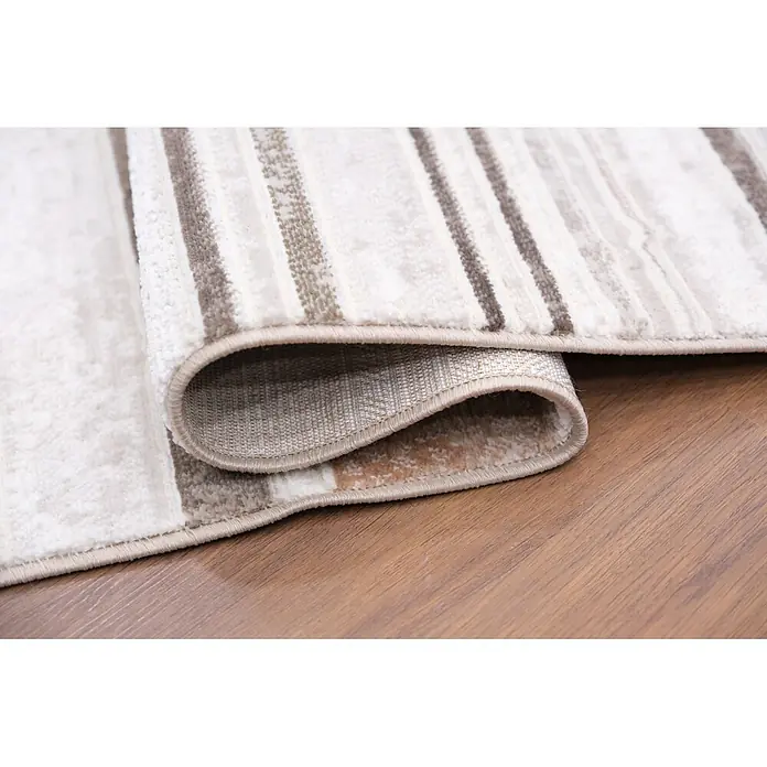 Alfombra de pelo corto Box - Natural, 230 x 160 cm, 100% poliéster - Una alfombra de rayas beige y marrón, parcialmente enrollada y de textura suave, sobre un suelo de madera.