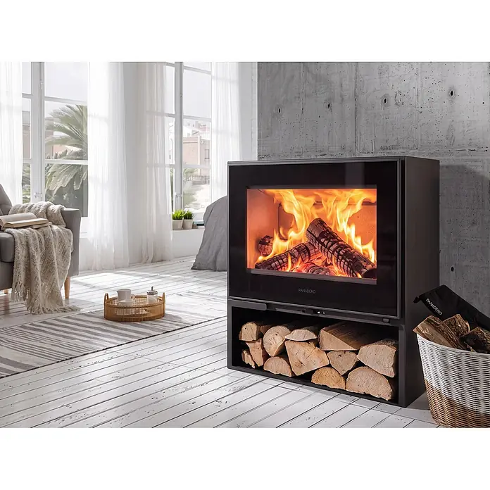 Panadero Kaminofen Borneo-S - 6,7 kW, Raumheizvermögen: 210 m³, Anthrazit - Moderner schwarzer Kaminofen mit Glastür, brennenden Holzscheiten und einem unteren Fach voller Brennholz in einem hellen Raum.