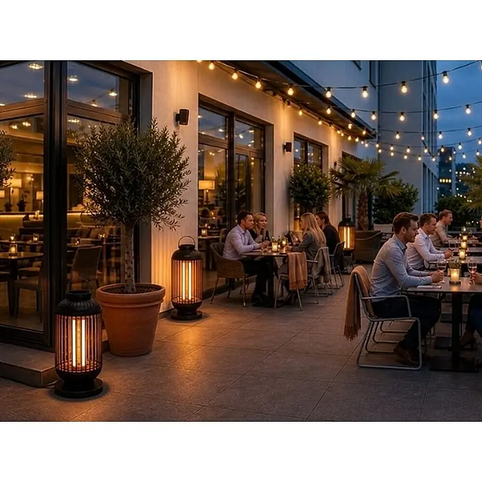 WDH Heizlüfter Standheizstrahler WDH-12HL Heizlaterne - 1,2 kW, Schwarz, IP54 - Schwarze Lamellen-Outdoor-Lampe mit vertikalem Warmlicht, die eine belebte Restaurantterrasse bei Dämmerung erhellt.