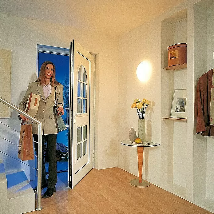 Steinel Plafón de techo y pared con sensor RS 16 L - 60 W, L x An x Al: 27,5 x 27,5 x 9,5 cm, Blanco, E27 - Una mujer entra por una puerta exterior blanca y parcialmente acristalada a un recibidor moderno, llevando bolsas.