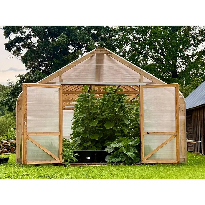 Garten Dream Gewächshaus Classic Wood - 450 x 800 cm, Material Seitenwand: Kunststoff, 10 mm, Braun - Ein Gewächshaus mit Holzrahmen, lichtdurchlässigen Platten und offenen Türen, gefüllt mit Pflanzen, auf einer Wiese.