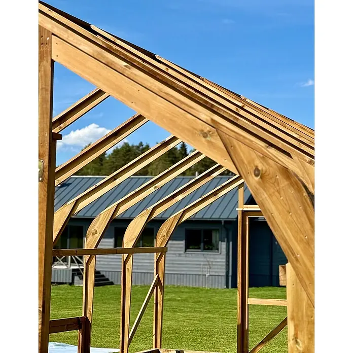 Garten Dream Gewächshaus Classic Wood - 450 x 800 cm, Material Seitenwand: Kunststoff, 10 mm, Braun - Heller Holzrahmen einer Gewächshaus- oder Gartenstruktur, vor blauem Himmel und entferntem Haus.