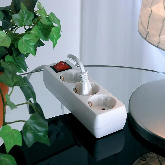 Steckdosenleiste - Anzahl Steckdosen Schuko: 3 Stk., Weiß, Kabellänge: 1,4 m, Mit Schalter - Nahaufnahme einer weißen Steckdosenleiste mit einem eingesteckten Stecker auf einem Tisch, neben einer Pflanze und einer Lampe.