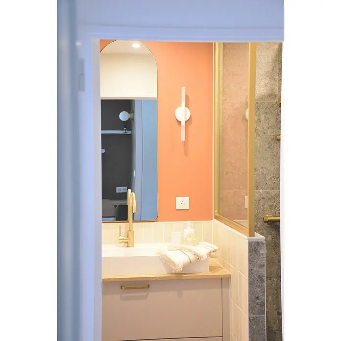 Hudor Waschtischarmatur Scamandre 6565 - Seidenmatt - Goldener Wasserhahn auf einem weißen Waschbecken mit hellem Unterschrank in modernem Bad mit oranger Wand und Dusche.