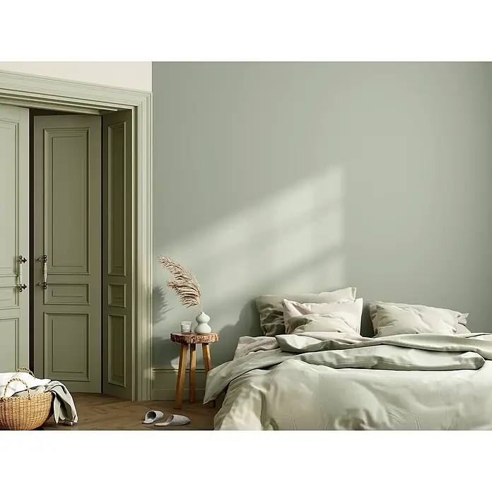 Caleo Color Wandfarbe Lehmfarbe Jade - Jade, Matt, 10 l - Gemütliches Schlafzimmer mit sanften grünen Wänden, passenden Türen und Bettwäsche in Hellgrün und Beige. Ein Holzhocker mit Vase steht am Bett.