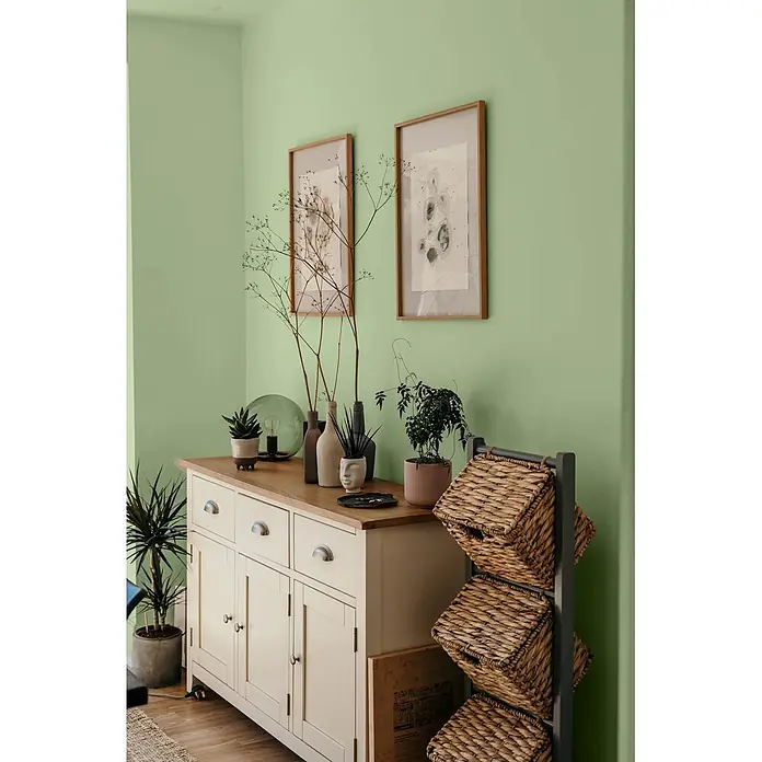 Caleo Color Wandfarbe Lehmfarbe Salbeiduft - Salbeiduft, Matt, 5 l - Helle Holzkommode mit Naturholzplatte, dekoriert mit Pflanzen und Kunst, neben einem Regal mit Korbtaschen.