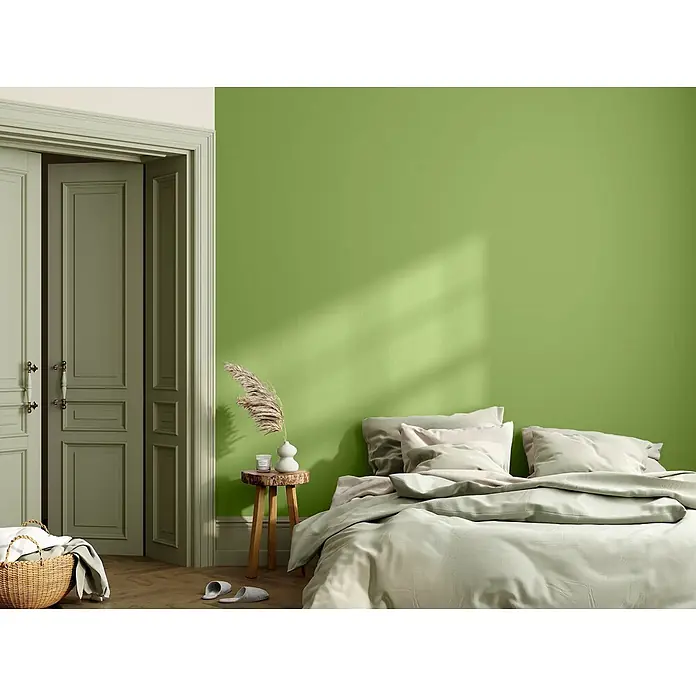 Caleo Color Wandfarbe Lehmfarbe Gingko - Gingko, Matt, 10 l - Schlafzimmerwand in Salbeigrün gestrichen, mit heller Bettwäsche und einem Holzhocker.