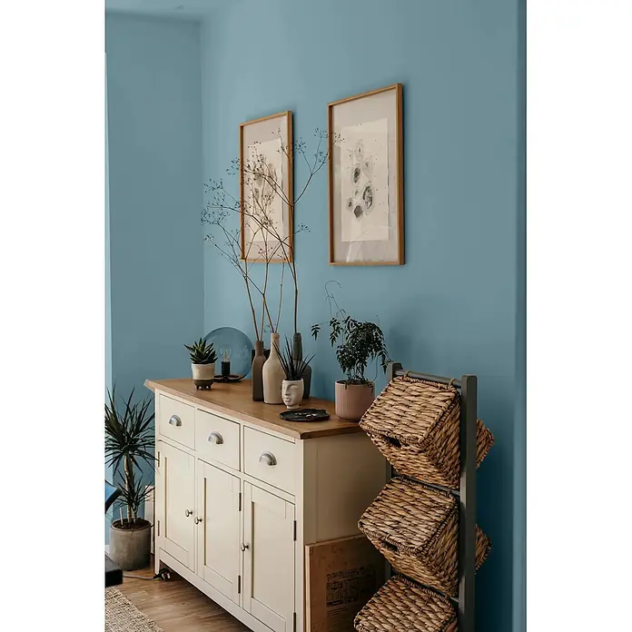 Caleo Color Wandfarbe Lehmfarbe Lagune - Lagune, Matt, 10 l - Cremefarbenes Holzbuffet mit Naturholzplatte in blauem Zimmer, daneben ein Regal mit Weidenkörben.