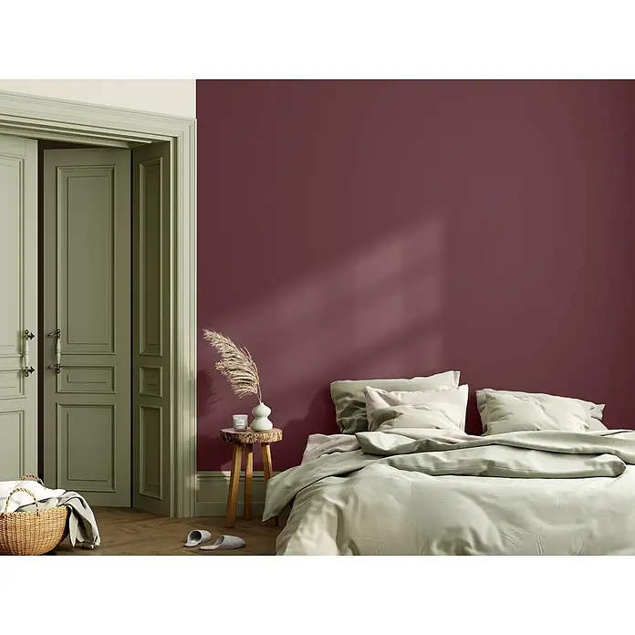 Caleo Color Wandfarbe Lehmfarbe Kaminabend - Kaminabend, Matt, 2,25 l - Hellgrüner Bettbezug und Kissen auf einem Bett vor einer dunkelroten Akzentwand.