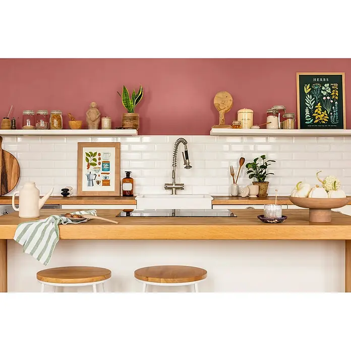 Caleo Color Wandfarbe Lehmfarbe Rosenknospe - Rosenknospe, Matt, 5 l - Eine lebendige Küche mit rosa Wand, weißen Fliesen und Holztheke mit Regalen. Dekoriert mit Küchenutensilien, Pflanzen und Kunst über zwei Hockern.