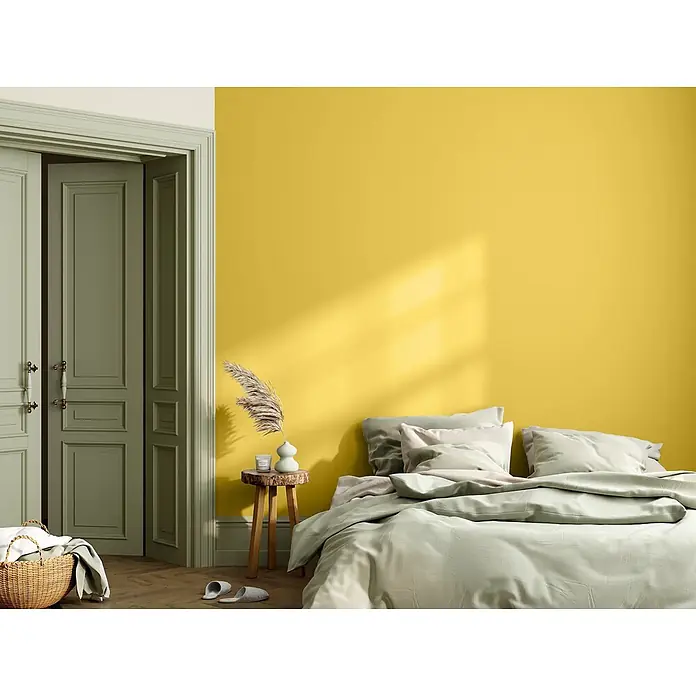 Caleo Color Wandfarbe Lehmfarbe Steppengras - Steppengras, Matt, 5 l - Ein Bett mit weicher salbeigrüner Bettwäsche in einem sonnigen Schlafzimmer mit leuchtend gelben Wänden und olivgrünen Türen.