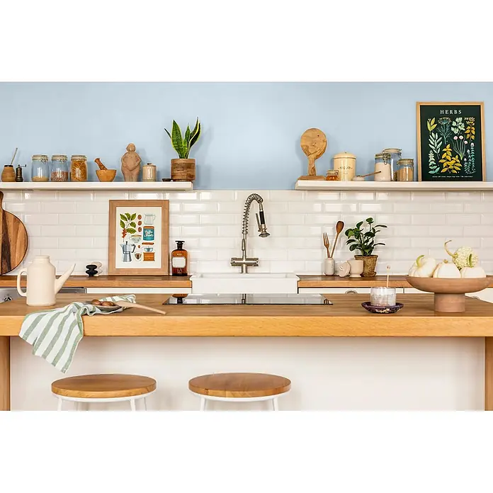 Caleo Color Wandfarbe Lehmfarbe Morgentau - Morgentau, Matt, 2,25 l - Eine gemütliche Küche mit hellblauen Wänden, weißen Fliesen und Holzregalen. Eine weiße Spüle und moderne Armatur sind in die Holztheke eingelassen, umgeben von Deko.