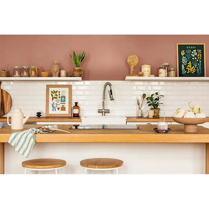 Caleo Color Wandfarbe Lehmfarbe Herbstlaub - Herbstlaub, Matt, 2,25 l - Eine warme, organisierte Küche mit Holztheke, weißem Fliesenspiegel und altrosafarbener Wand. Regale zeigen Gläser und Pflanzen, schaffen eine einladende Atmosphäre.