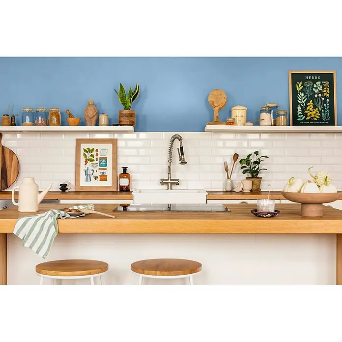 Caleo Color Wandfarbe Lehmfarbe Sommerregen - Sommerregen, Matt, 5 l - Eine gemütliche Küche mit hellblauen Wänden, weißen Fliesen, Arbeitsplatten aus Holz und offenen Regalen, mit Kochutensilien und Dekoration.