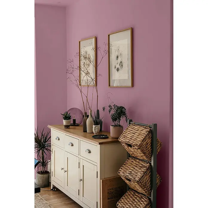 Caleo Color Wandfarbe Lehmfarbe Königsflieder - Königsflieder, Matt, 5 l - Ein cremefarbenes Sideboard mit Holzplatte und ein dunkles Regal mit gewebten Weidenkörben in einem Raum mit lavendelfarbenen Wänden.