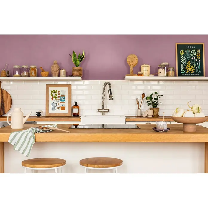 Caleo Color Wandfarbe Lehmfarbe Königsflieder - Königsflieder, Matt, 5 l - Eine gemütliche Küche mit lila Wänden, weißen Fliesen und Holztheken, mit offenen Regalen voller Küchenartikel und Dekoration.
