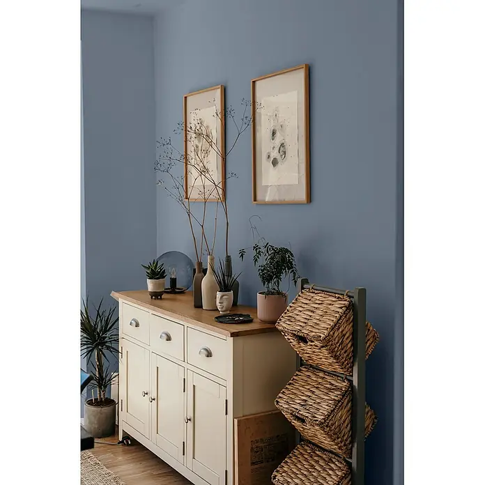 Caleo Color Wandfarbe Lehmfarbe Wolkenbett - Wolkenbett, Matt, 0,15 l - Helles cremefarbenes Holzbuffet mit Holzplatte und Metallgriffen, dekoriert mit Pflanzen, neben einem Regal mit geflochtenen Körben in einem blau gestrichenen Raum.