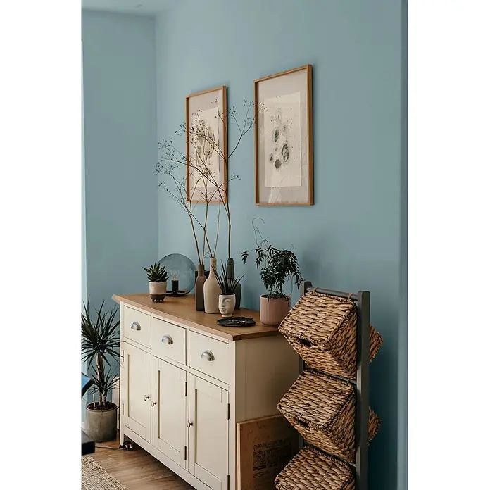 Caleo Color Wandfarbe Lehmfarbe Traumsand - Traumsand, Matt, 2,25 l - Ein cremefarbenes Sideboard aus Holz mit naturfarbener Holzplatte und Metallgriffen, ergänzt durch ein graues Regal mit drei geflochtenen Körben.