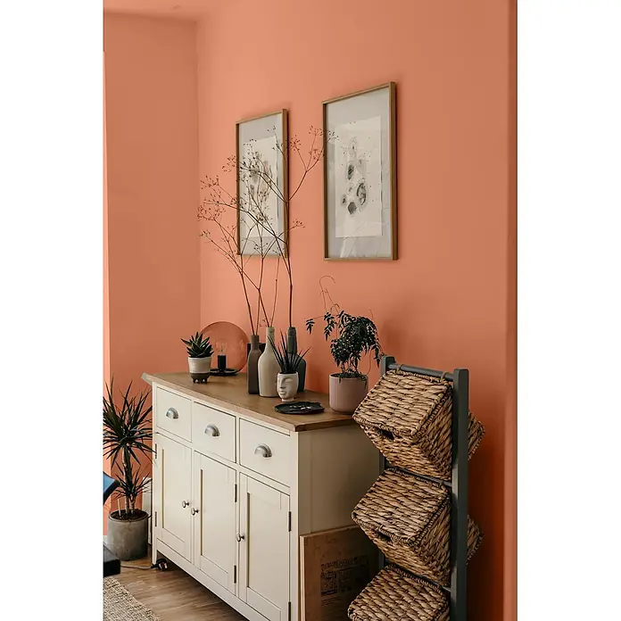 Caleo Color Wandfarbe Lehmfarbe Tonscherbe - Tonscherbe, Matt, 2,25 l - Ein Zimmer mit warmen pfirsichfarbenen Wänden, einem cremefarbenen Sideboard und einem dunkelgrauen Regal mit Weidenkörben.