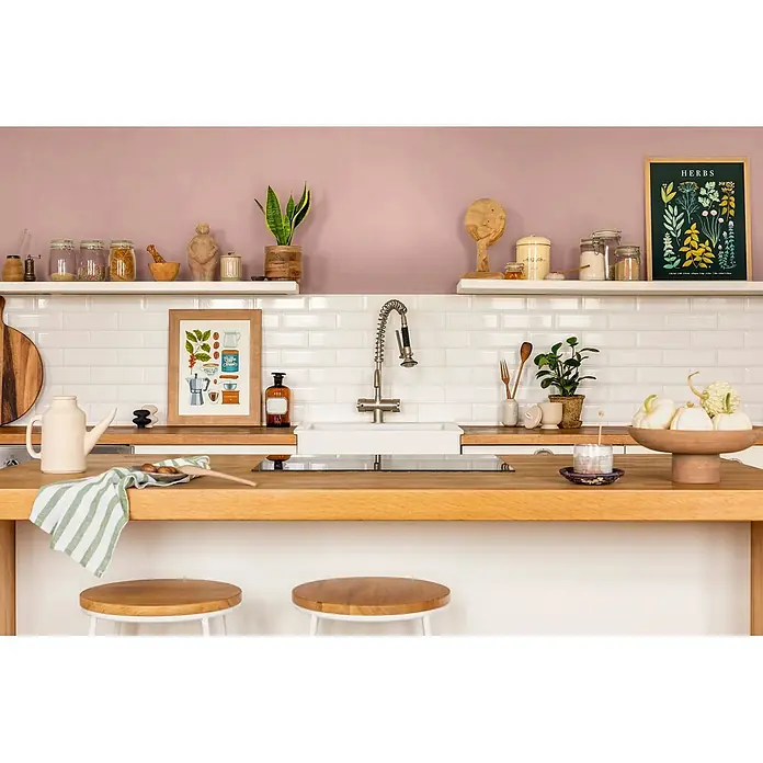 Caleo Color Wandfarbe Lehmfarbe Magnolie - Magnolie, Matt, 0,15 l - Eine moderne Küche mit hellen Holz-Arbeitsplatten, weißen Metrofliesen und Regalen mit Pflanzen, Gewürzen und Bildern. Zwei Holzbarhocker vervollständigen das gemütliche Ambiente.
