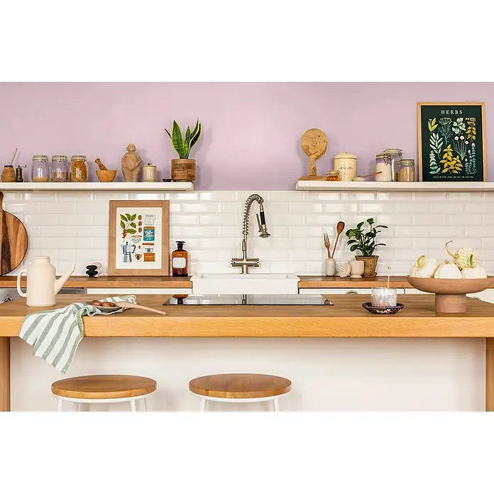 Caleo Color Wandfarbe Lehmfarbe Blütenzauber - Blütenzauber, Matt, 5 l - Eine helle, gemütliche Küche mit Holzarbeitsplatten, weißen Metrofliesen, einer hellvioletten Wand und offenen Regalen mit Pflanzen und Gläsern.