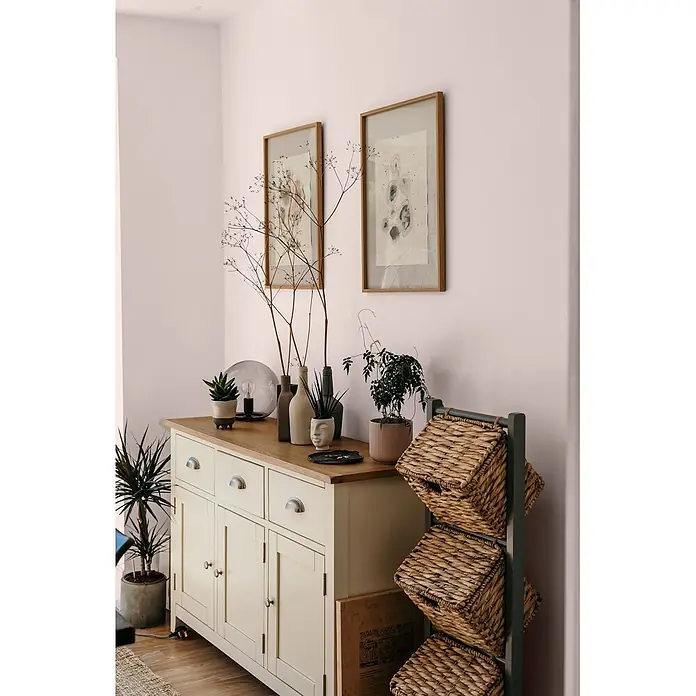 Caleo Color Wandfarbe Lehmfarbe Blütenpracht - Blütenpracht, Matt, 0,15 l - Cremeweiße Holzkommode mit Holzplatte, Metallgriffen und Pflanzen. Daneben ein dunkles Regal mit geflochtenen Körben.