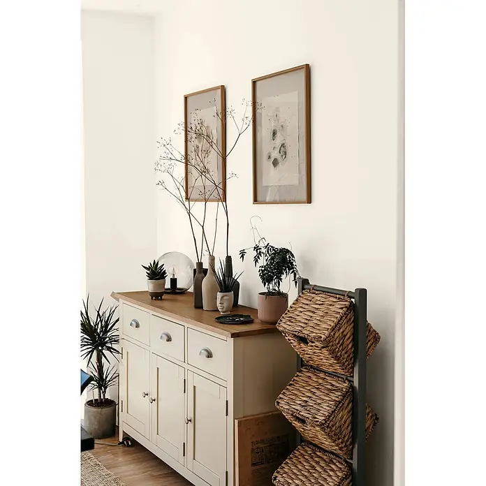 Caleo Color Wandfarbe Lehmfarbe Lammwolle - Lammwolle, Matt, 2,25 l - Cremefarbenes Sideboard mit Holzplatte und Pflanzen, daneben ein dunkelgraues Regal mit geflochtenen Weidenkörben.