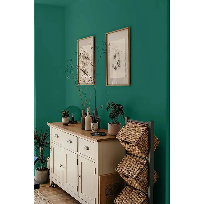 Caleo Color Wandfarbe Lehmfarbe Palmenblatt - Palmenblatt, Matt, 2,25 l - Cremefarbenes Holzbuffet mit Naturholzplatte, daneben ein dunkelgraues Regal mit drei geflochtenen Weidenkörben, dekoriert mit Pflanzen und Vasen.