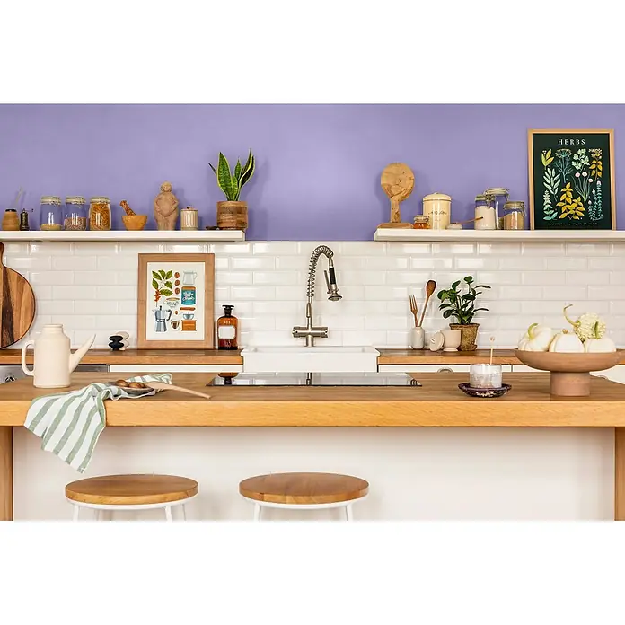 Caleo Color Wandfarbe Lehmfarbe Veilchenblüte - Veilchenblüte, Matt, 10 l - Moderne Küche mit hellen Holz-Arbeitsplatten, weißen Metro-Fliesen und offenen Regalen mit Gläsern, Pflanzen und Kunst. Spüle und Barhocker sind integriert.