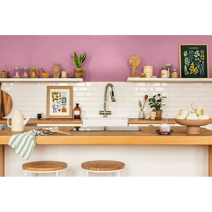 Caleo Color Wandfarbe Lehmfarbe Himbeere - Himbeere, Matt, 2,25 l - Helle, stilvolle Küche mit Holztheke, weißen Metrofliesen und lila Wand, mit Pflanzen und Küchenaccessoires.