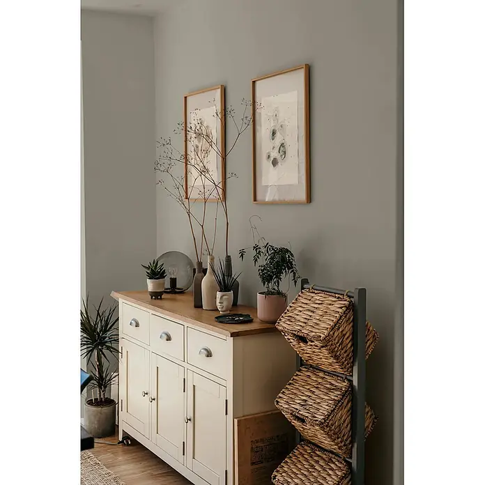 Caleo Color Wandfarbe Lehmfarbe Mangrovenwald - Mangrovenwald, Matt, 2,25 l - Cremefarbenes Sideboard mit heller Holzplatte und silbernen Griffen, drei Schubladen und drei Schränken, in einem dekorierten Wohnraum.