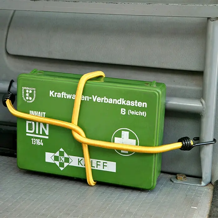UniTEC Gepäckspanner - Länge: 100 cm, 2 Haken - Ein grüner Verbandskasten für Kraftwagen ist mit einem gelben Gummiband an einer Metallstange befestigt. DIN 13164 ist aufgedruckt.