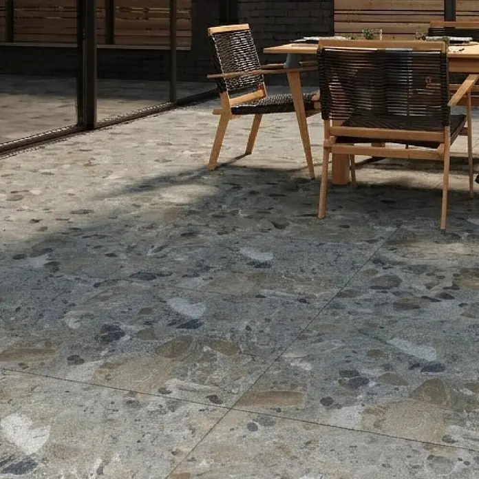 Feinsteinzeugfliese Vogue - 60 x 60 cm, Grey, Matt - Terrassenfliesen in Terrazzo-Optik, Grautöne mit Naturstein-Aggregaten, unter einem Esstisch mit Stühlen.